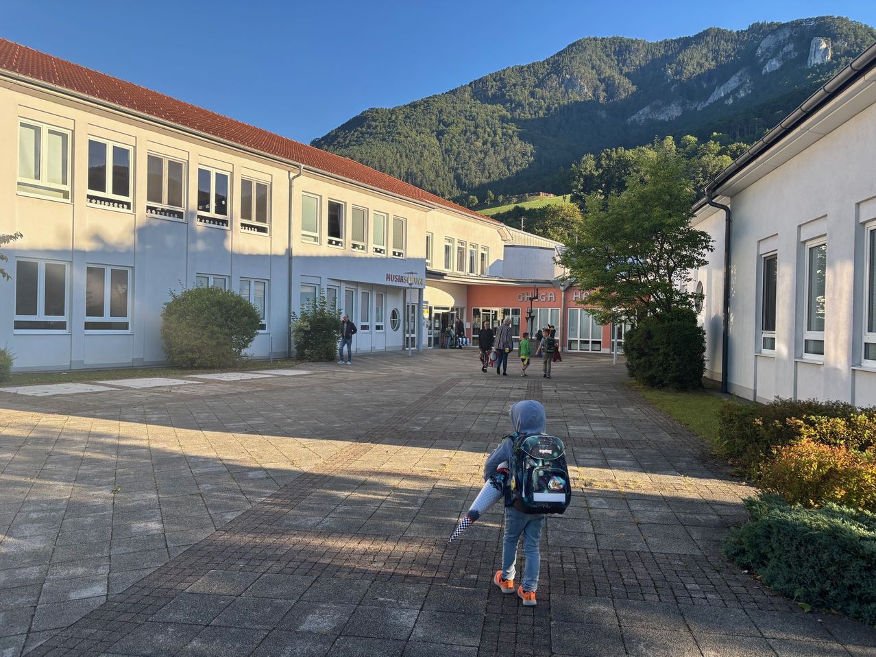 Ein Kind geht mit einem Rucksack durch einen Innenhof. Im Hintergrund befinden sich Gebäude und ein Berg. Mehrere Menschen gehen in Richtung der Gebäude.