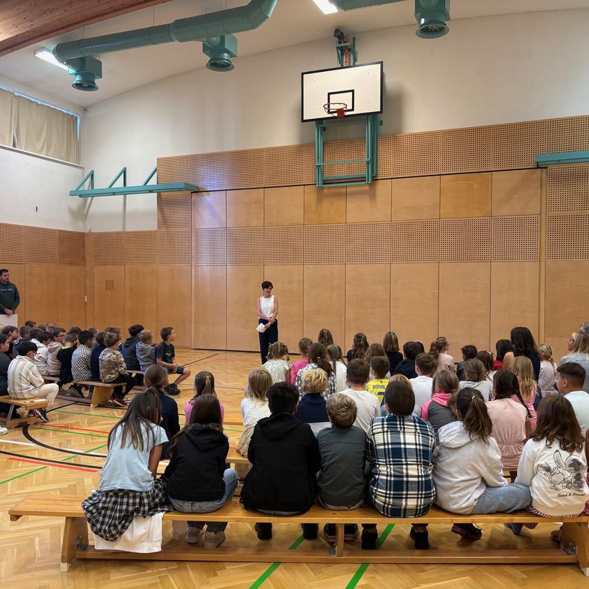Eine Gruppe von Schülern sitzt auf Holzbänken in einer Turnhalle und hört einer Frau zu, die vor einem Basketballkorb steht. Ein Mann steht in der Ecke, und die Halle hat Holzwände und eine Decke mit Lichtern.