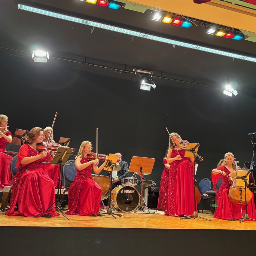 Eine Gruppe von Frauen in roten Kleidern spielt Violinen auf der Bühne, während ein Mann in Anzug die Drums spielt. Scheinwerfer sind an der Decke.