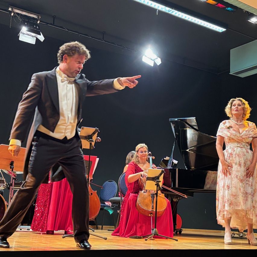 Ein Mann in einem schwarzen Anzug zeigt auf eine Frau, die Cello spielt, während eine Frau in einem Blumenkleid zuschaut. Ein Pianist ist ebenfalls auf der Bühne mit ihnen.