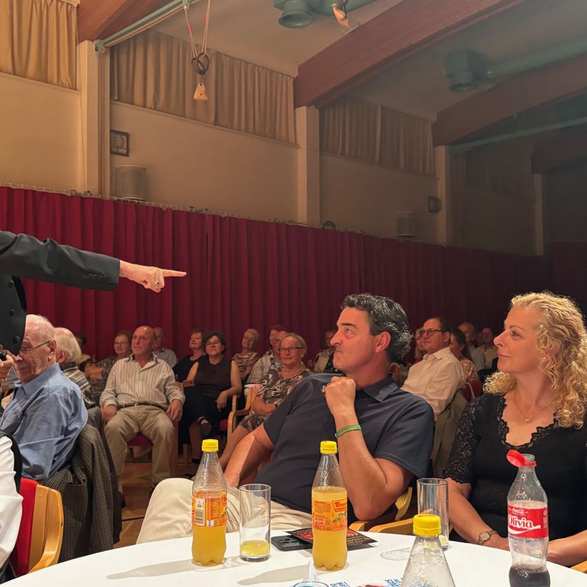 Ein Mann zeigt auf ein sitzendes Publikum in einem Theater. Flaschen mit Orangensaft und eine Coca-Cola-Flasche stehen auf dem Tisch vor einem Mann und einer Frau.