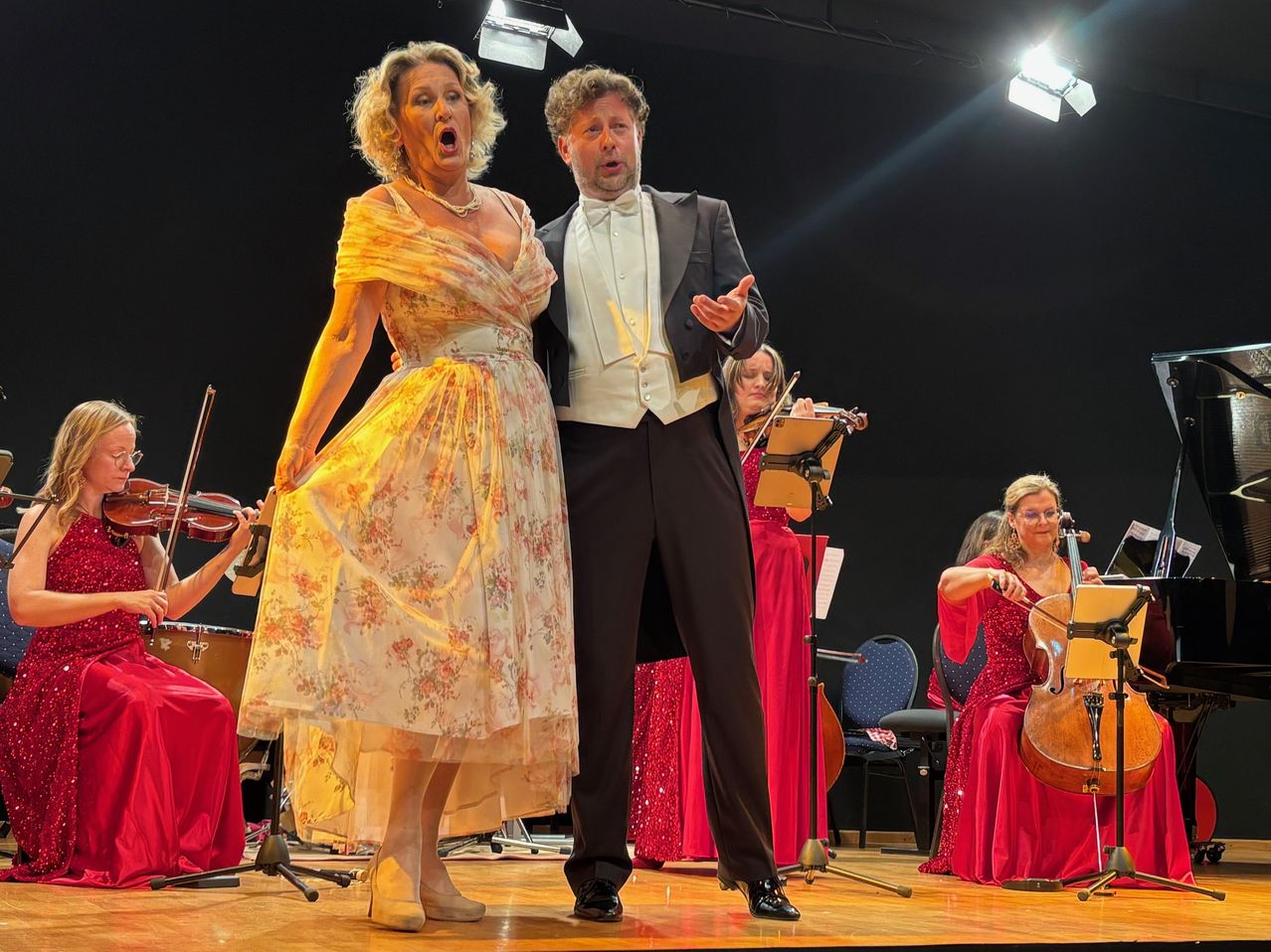 Eine Frau und ein Mann stehen auf der Bühne, beide leger gekleidet. Sie singen und scheinen ein Duett darzubieten. Hinter ihnen spielen Musiker verschiedene Instrumente, darunter Geige und Cello.