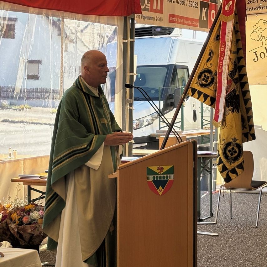 Ein Priester steht an einem Podium mit einem Mikrofon vor einer Fahne. Dahinter ist ein Lieferwagen geparkt und ein Gebäude mit Zaun ist sichtbar.