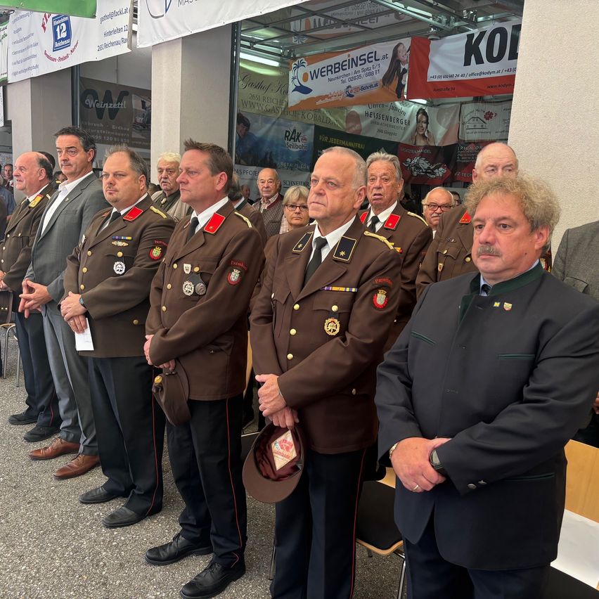 Eine Gruppe von Militäroffizieren, gekleidet in braune Uniformen, steht in einer Reihe. Einer hält einen Hut, während andere Dokumente in der Hand haben. Ein Mann in einem schwarzen Anzug steht vor ihnen.