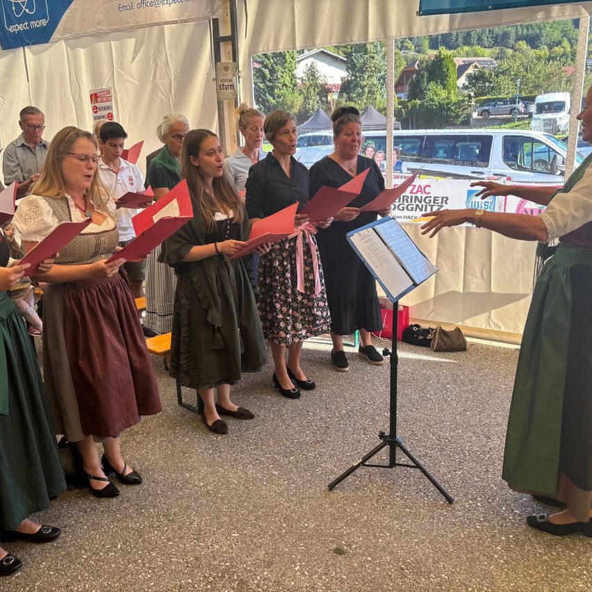 Ein Chor probt in einem Zelt. Eine Frau singt und dirigiert sie. Sie halten rote Notenblätter. Dahinter ist ein verschwommener Blick auf ein weißes Zelt und Autos, die vor einem Haus geparkt sind.