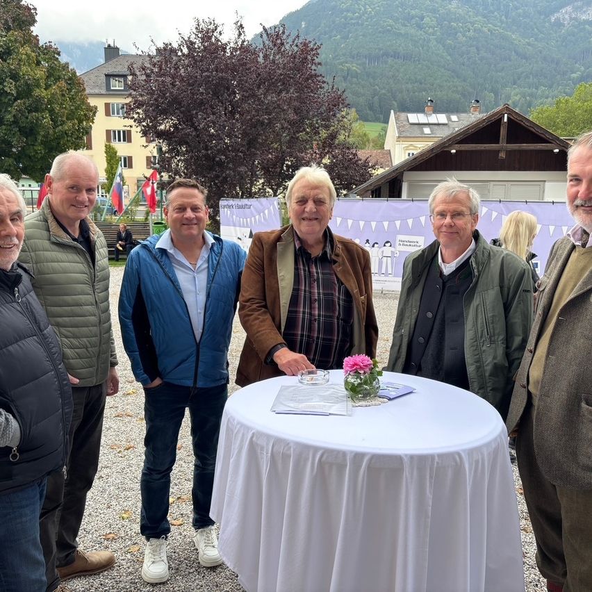 Fünf Männer stehen lächelnd um einen Tisch herum. Dahinter ein dekorativer Hintergrund und ein Gebäude mit Bergen im Hintergrund.