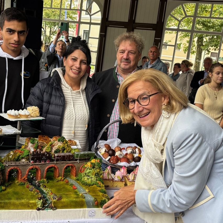 Eine Gruppe von Menschen versammelt sich um einen Tisch mit Kuchen und Desserts, eine Frau lächelt. Hinter ihnen machen andere Fotos. Draußen gibt es Bäume und Gebäude.