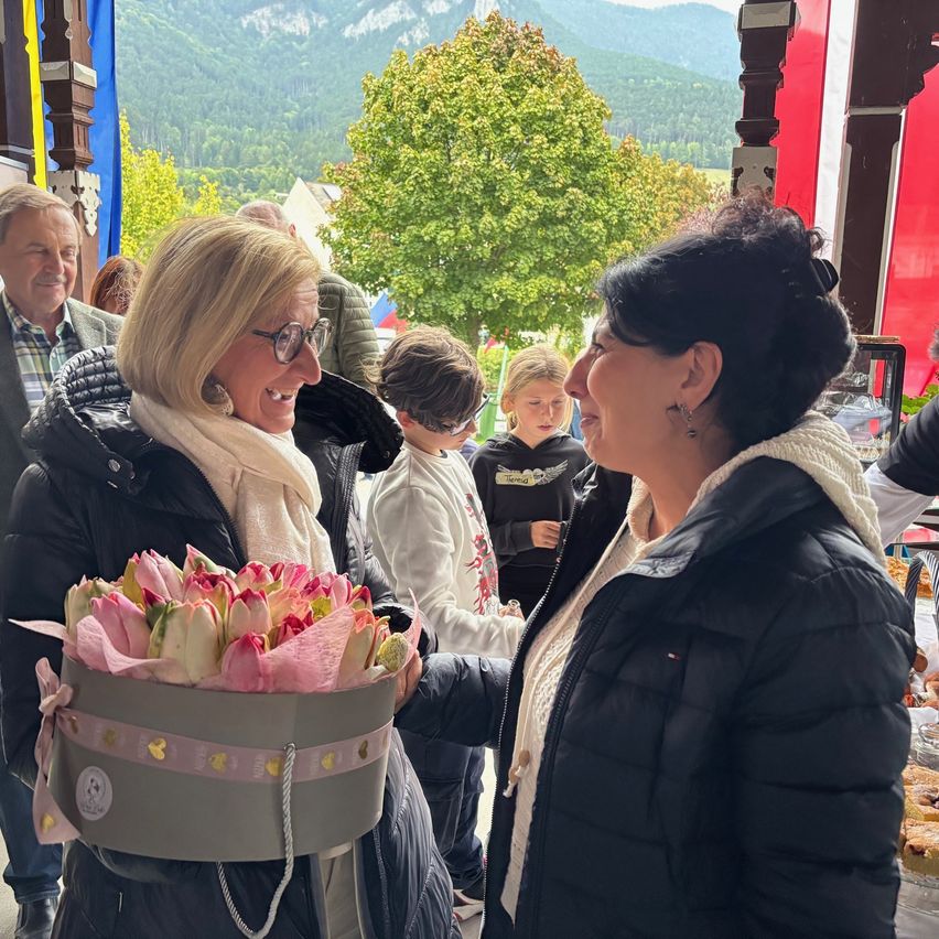 Zwei Frauen lächeln sich an, eine hält einen Strauß pinker Blumen. Im Hintergrund steht eine Gruppe von Menschen in der Nähe eines Tisches mit Essen, Berge in der Ferne.