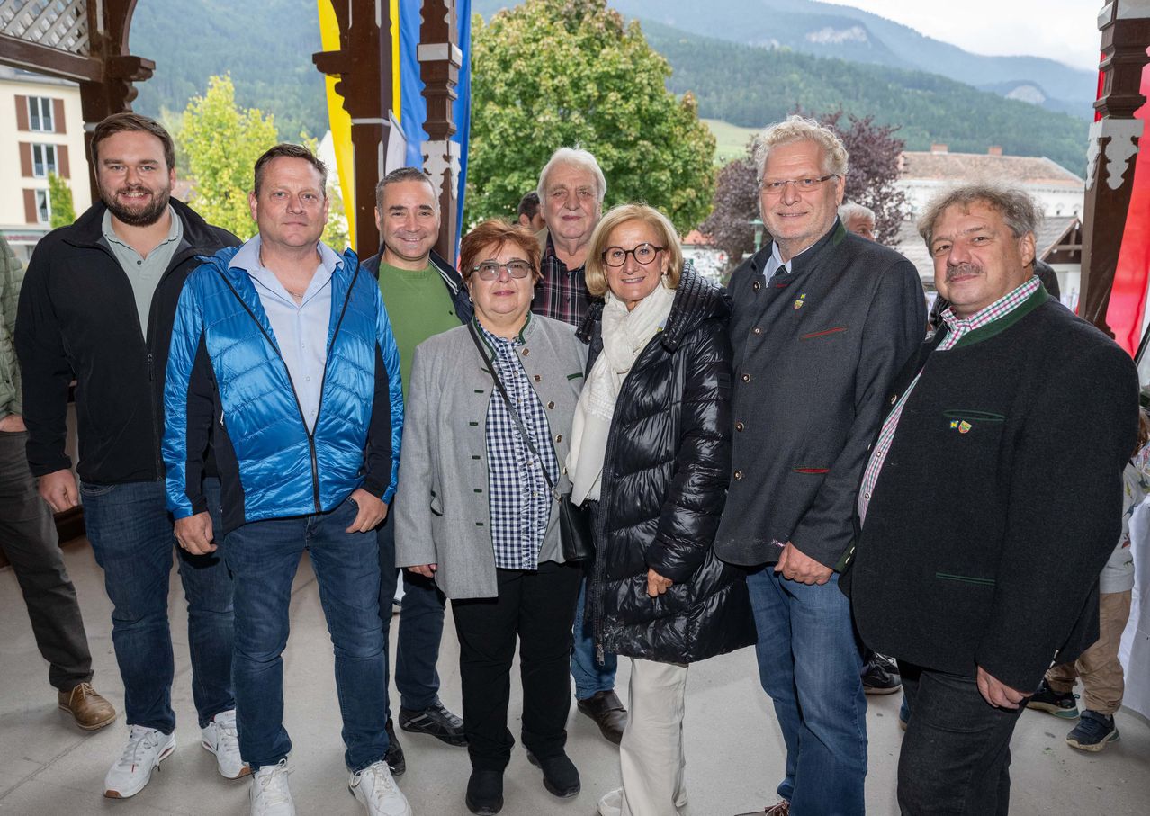 Eine Gruppe von Menschen, die für kühleres Wetter gekleidet sind, steht zusammen auf einer Veranda mit Bergen im Hintergrund. Sie scheinen für ein Foto zu posieren.