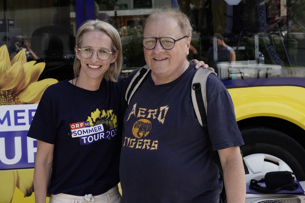 Ein Mann und eine Frau stehen lächelnd vor einem Bus. Sie tragen passende blaue T-Shirts. Das Shirt der Frau hat eine Sonnenblume und den Text 'Sommer Tour 2021'. Das Shirt des Mannes zeigt 'Aero Tigers' mit einem Tiger-Grafik.