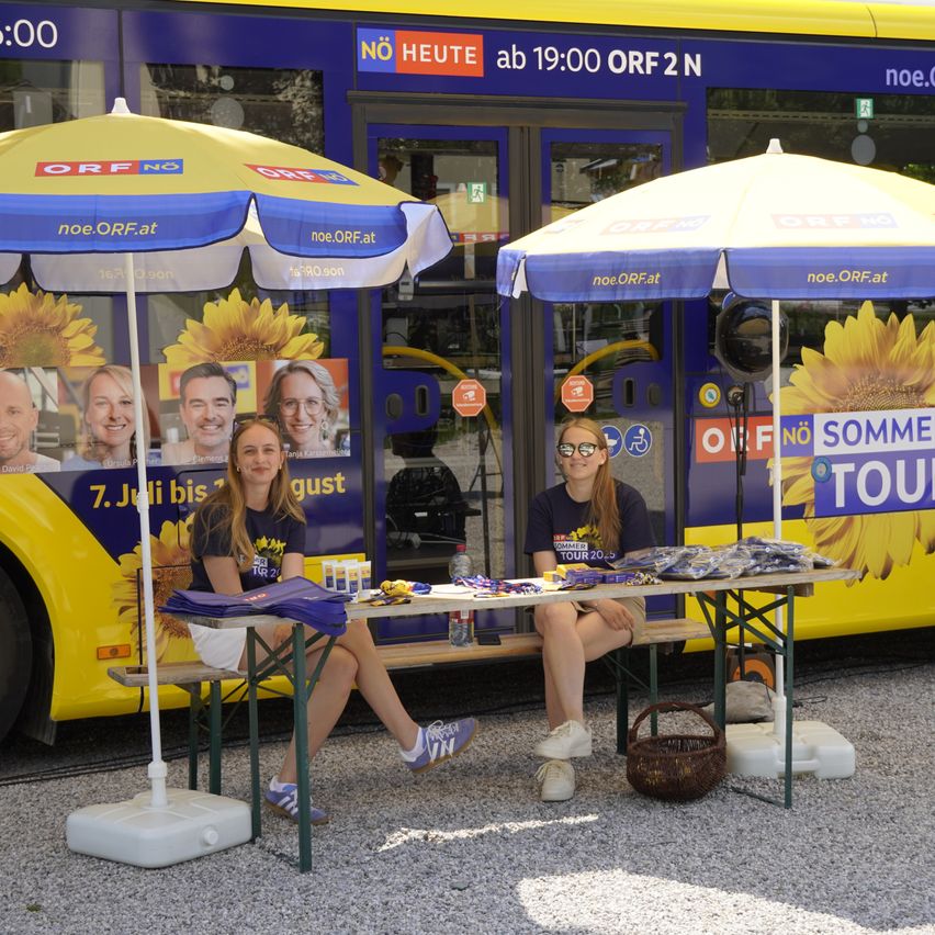 Zwei Frauen sitzen unter Sonnenschirmen an einem Tisch, mit einem Bus dahinter. Der Bus hat Werbung für den ORF2N-Kanal.