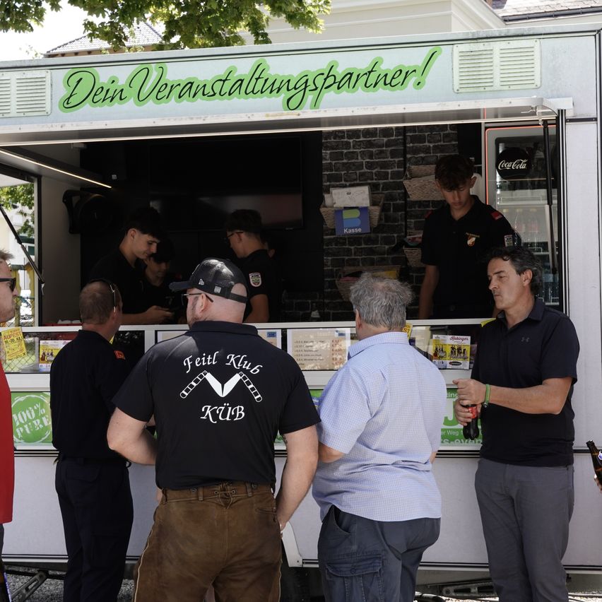 Ein Imbisswagen mit dem Schild 'Dein Veranstaltungspartner!' steht offen und serviert Essen. Zwei Köche sind drinnen, und mehrere Leute stehen draußen, darunter ein Mann in einem schwarzen Shirt mit weißem Text.