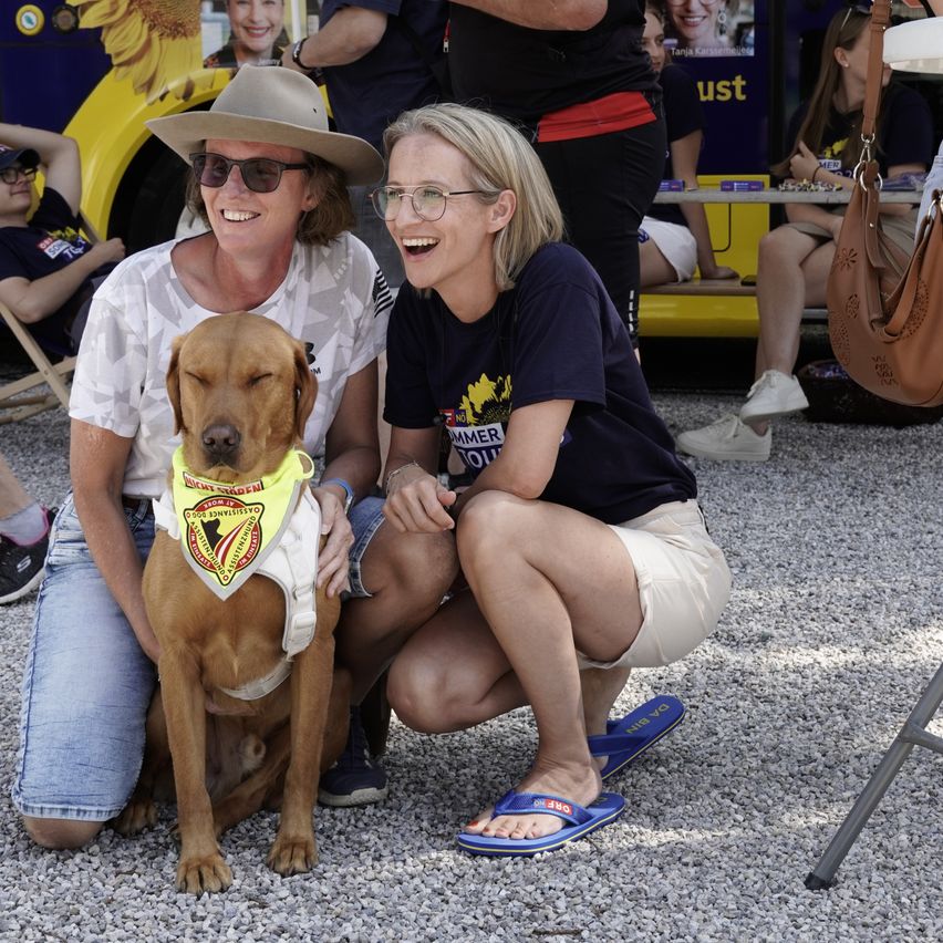 Zwei Frauen mit einem Hund. Die Frau auf der linken Seite trägt eine Mütze, eine Sonnenbrille und ein Camouflage-Shirt. Die Frau auf der rechten Seite trägt eine Brille, ein schwarzes Shirt und blaue Flip-Flops. Der Hund hat ein gelbes Halsband. Hinter ihnen sitzen Leute auf einer Bank.
