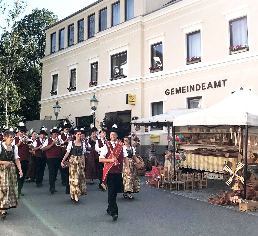 Eine Gruppe von Menschen marschiert spielend Instrumente die Straße hinunter, vor einem Gebäude mit der Aufschrift Gemeindeamt.