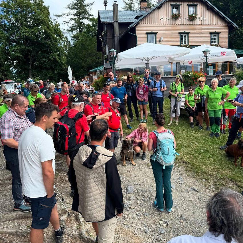 Eine Gruppe von Wanderern in grünen Shirts versammelt sich vor einem Haus mit Schirmen, einige mit Rucksäcken und Hunden, vor Bäumen und einem Rasen.