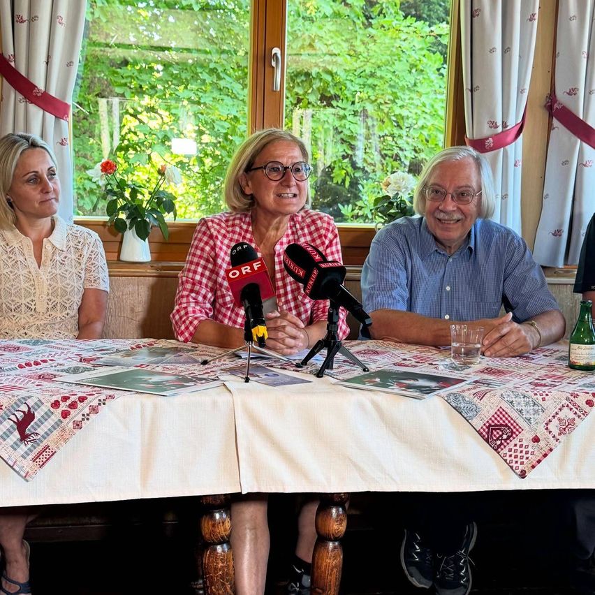 Drei Personen sitzen an einem Tisch mit Mikrofonen. Eine Frau trägt eine Brille und ein rot-weiß kariertes Hemd. Neben ihr sitzt ein Mann in einem blauen Hemd, der lächelt. Hinter ihnen zeigt ein Fenster einen Blick auf Bäume und eine Vase mit Blumen.