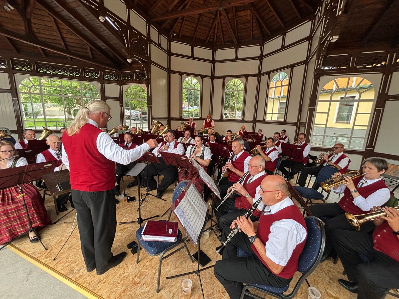 Eine Gruppe von Musikern in roten Westen und weißen Hemden spielt Instrumente in einem Pavillon mit Holzdecke und großen Fenstern.