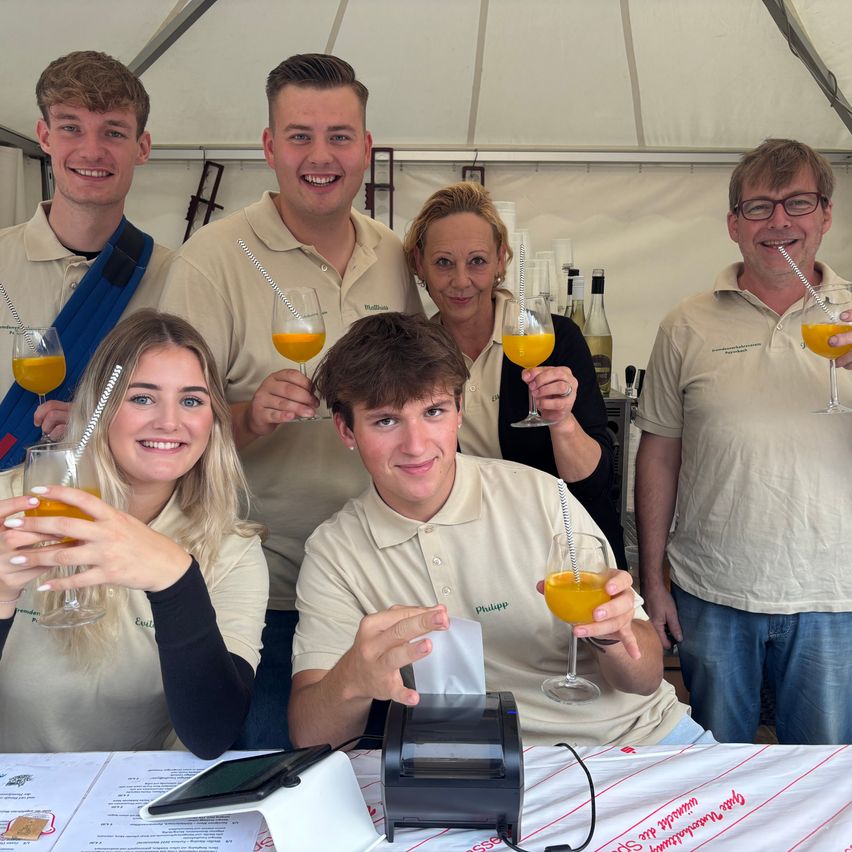 Eine Gruppe junger Menschen steht in einem Zelt und hält orangefarbene Getränke in Weingläsern. Sie lächeln und posieren für ein Foto. Der Tisch vor ihnen hat eine Kasse und ein Tablet.
