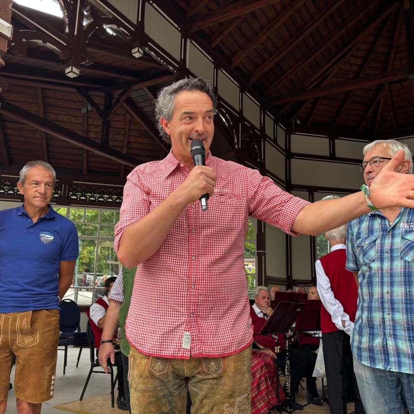 Ein Mann spricht in ein Mikrofon in einem Pavillon, trägt ein rotes kariertes Hemd und braune Hosen, während zwei Männer hinter ihm stehen, einer in einem blauen Shirt und der andere in einem karierten Shirt.