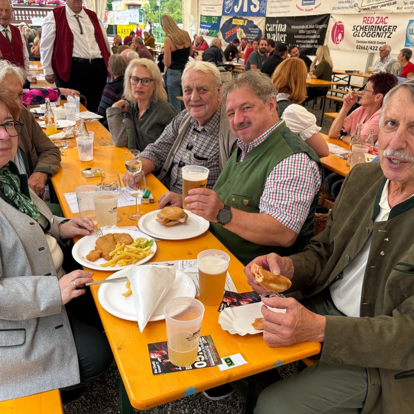 Eine Gruppe von Menschen sitzt an einem langen Tisch und genießt Essen und Getränke. Sie scheinen sich in einer festlichen Umgebung zu befinden, möglicherweise in einem Biergarten.