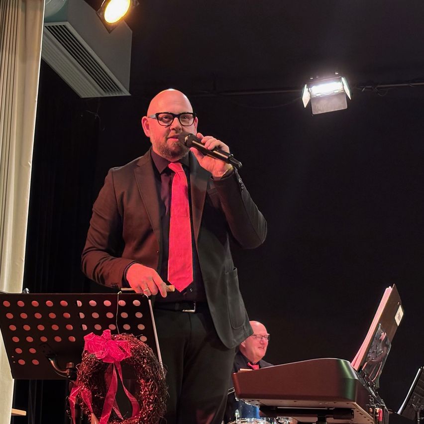 Ein Mann in einer roten Krawatte und mit Brille steht auf der Bühne und hält ein Mikrofon. Hinter ihm spielt ein anderer Mann Klavier. Vor ihm steht ein Notenständer mit einem Kranz. Scheinwerfer sind an der Decke montiert.