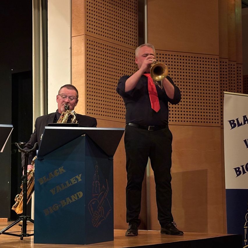 Zwei Männer spielen auf der Bühne mit einem Schild, das 'Black Valley Big Band' steht. Einer spielt Saxophon, der andere Trompete. Hinter ihnen ist eine braune Wand mit Perforationen.