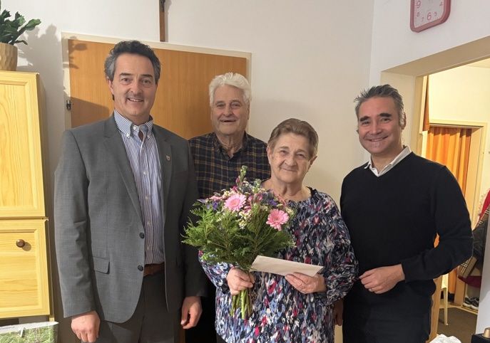 Drei Männer und eine Frau stehen in einem Raum mit Blumen und einem Schrank und lächeln für ein Foto.
