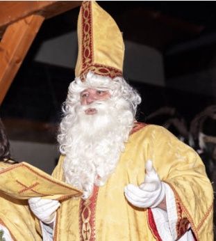 Ein Mann, der als Weihnachtsmann gekleidet ist, hält ein Buch in der Hand und gibt einen Daumen hoch, steht vor einer Holzstruktur.