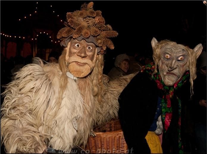 Zwei Personen in aufwendigen Kostümen mit Masken und Perücken stehen zusammen in einer Menschenmenge, eine hält einen Korb. Der Hintergrund zeigt festliche Lichter und eine verschwommene Menge.