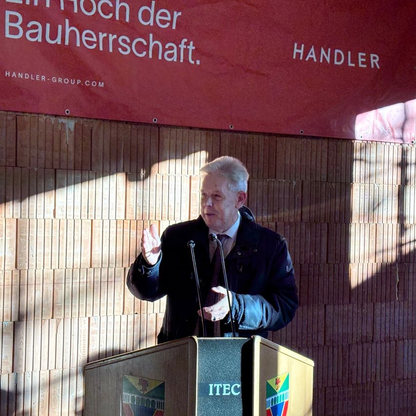 Ein älterer Mann in Anzug und Krawatte steht an einem Rednerpult mit Mikrofon und hält eine Rede vor einer Ziegelwand mit einem Schild, das 'Ein Hoch der Bauherrschaft' besagt.