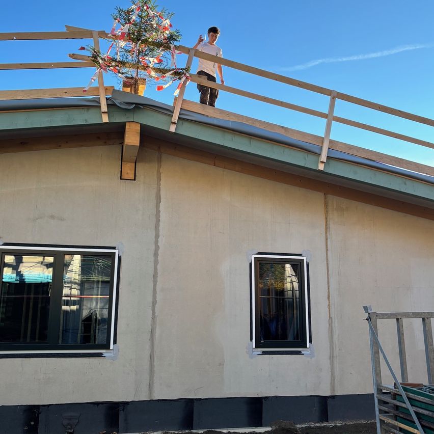 Eine Person steht auf dem Dach eines im Bau befindlichen Gebäudes, neben einem kleinen Weihnachtsbaum, an einem sonnigen Tag.