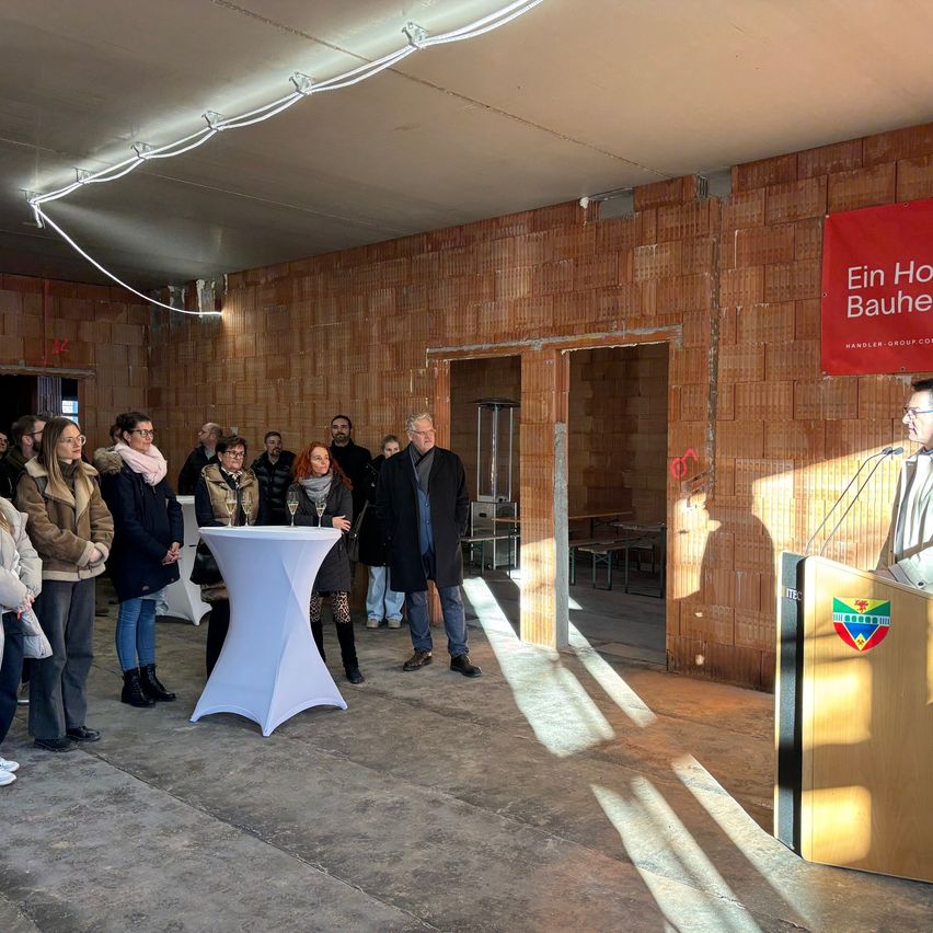Eine Gruppe von Menschen steht um einen Tisch und ein Podium herum, wobei ein Mann am Podium spricht. Ein roter Banner mit der Aufschrift 'Ein Haus Bauen' ist an einer Ziegelwand angebracht.
