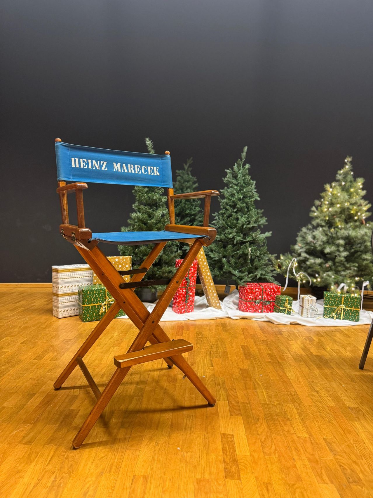 Ein Regiestuhl mit 'Heinz Marecek' darauf, auf einem Holzboden mit mehreren Weihnachtsbäumen und Geschenken darum.