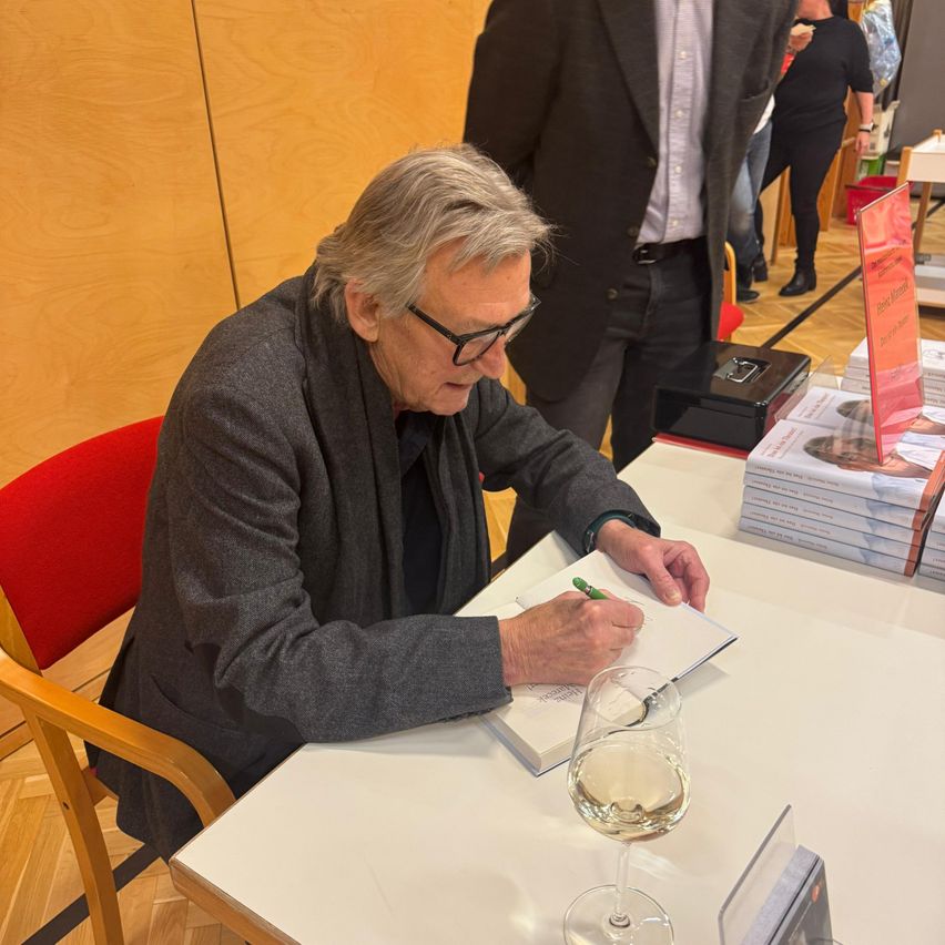 Ein älterer Mann mit Brille sitzt an einem Tisch und signiert Bücher, mit einem Glas Wein und Stapeln von Büchern vor ihm.