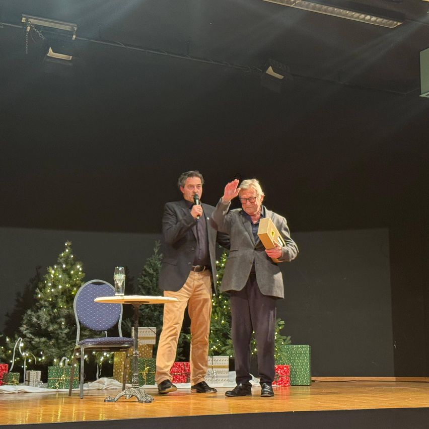 Zwei Männer stehen auf der Bühne, einer hält ein Mikrofon und der andere eine Box, sie lächeln und winken. Hinter ihnen stehen Weihnachtsbäume mit Geschenken. Ein Tisch mit einem Glas steht auf der linken Seite.