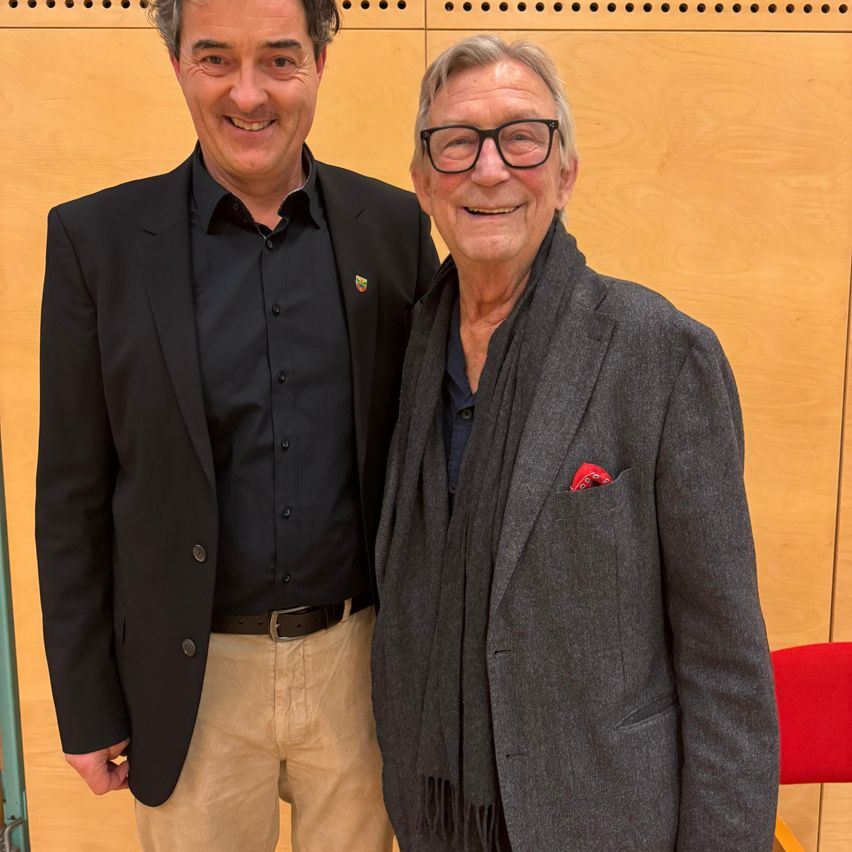 Zwei Männer stehen nebeneinander und lächeln. Einer trägt einen schwarzen Anzug, der andere einen grauen Mantel. Sie befinden sich in einem Raum mit einer Holzwand.