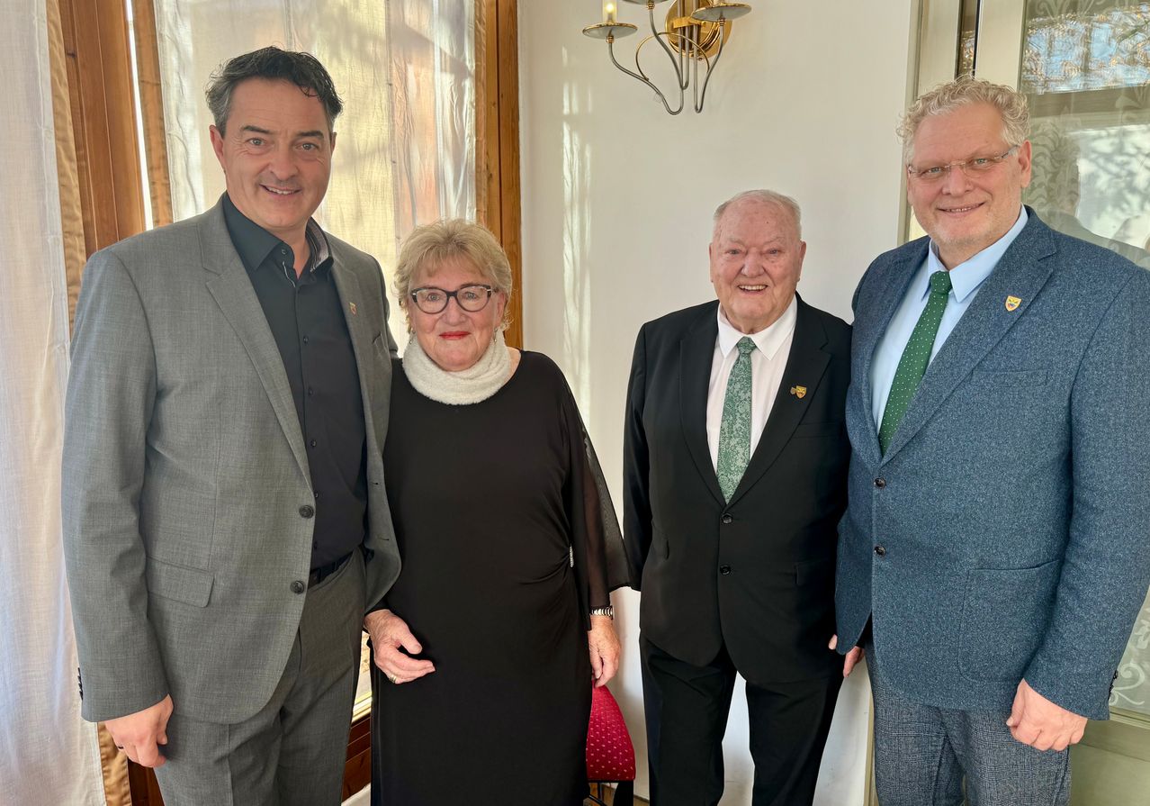 Vier Menschen stehen lächelnd zusammen, zwei Männer in Anzügen und eine Frau in einem Kleid. Der Mann links trägt einen grauen Anzug, die Frau ein schwarzes Kleid. Der Mann rechts trägt einen blauen Anzug.