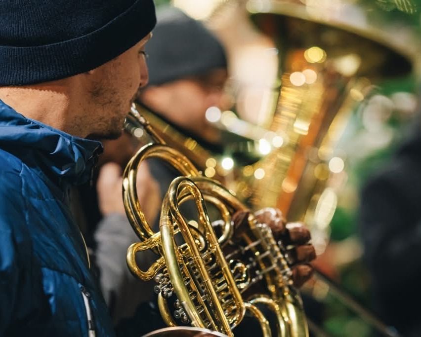 Ein Mann in einer blauen Jacke spielt eine französische Horn, umgeben von anderen Musikern in einer schwach beleuchteten Umgebung.