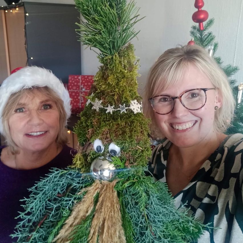 Zwei Frauen lächeln in einem Raum mit einem Weihnachtsbaum. Eine hält eine witzige Pflanzendekoration, die einem lächelnden Gesicht mit Augen und Nase ähnelt. Hinter ihnen steht ein geschmückter Weihnachtsbaum und verpackte Geschenke, die die festliche Atmosphäre verstärken.