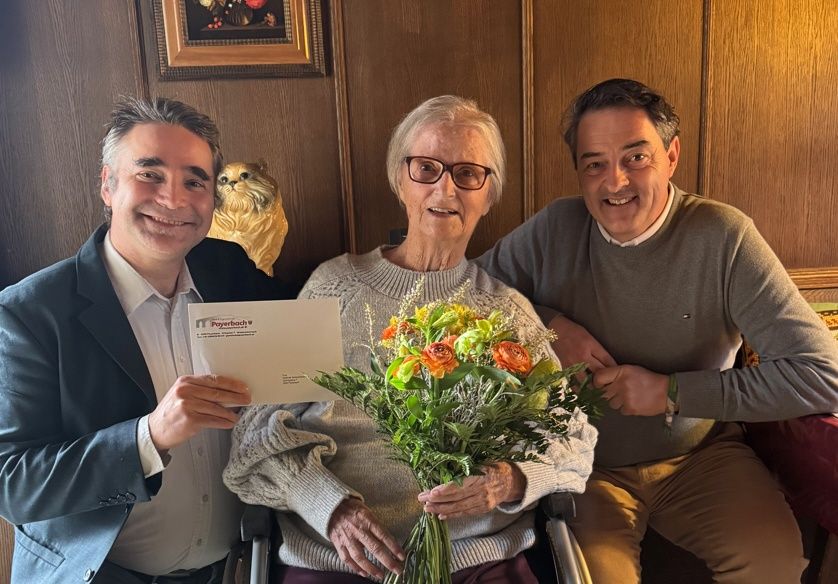 Drei Menschen lächeln, halten Blumen und ein Zertifikat. Die Frau im Rollstuhl trägt eine Brille. Hinter ihnen eine Holzwand mit einem gerahmten Bild.