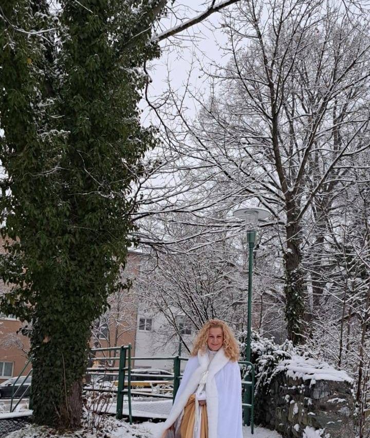 Eine Frau in einem weißen Mantel steht draußen und lächelt, mit schneebedeckten Bäumen und einem Laternenpfahl hinter ihr.