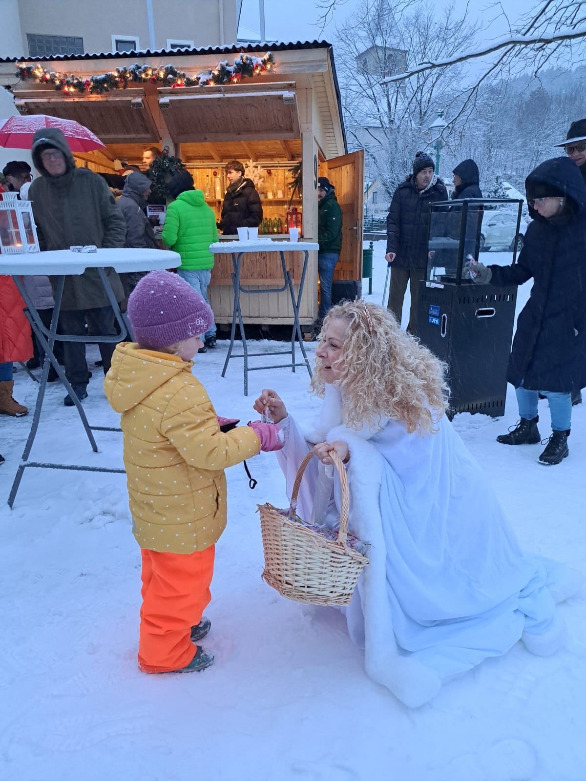 Eine als Prinzessin verkleidete Frau kniet im Schnee und gibt einem Kind in einer gelben Jacke einen Korb. Hinter ihnen versammeln sich Menschen in warmer Kleidung um einen Imbissstand.