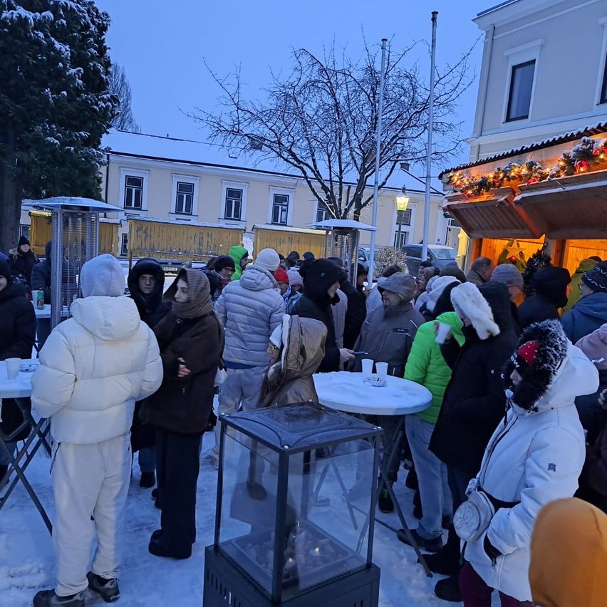 Eine Menschenmenge, alle warm gekleidet, versammelt sich draußen im Schnee, mit Tischen und einem Heizgerät im Vordergrund. Bäume und ein Gebäude mit festlichen Dekorationen sind im Hintergrund.