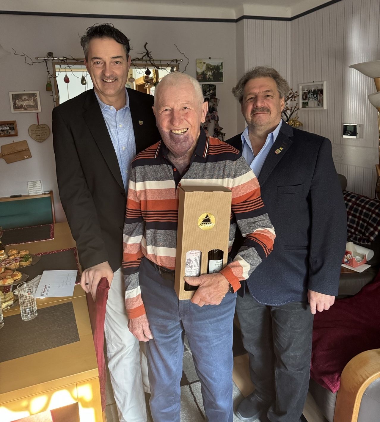 Drei Männer stehen eng beieinander in einem Raum. Der Mann in der Mitte hält eine Box mit Weinflaschen und lächelt. Dahinter steht ein Tisch mit Essen und eine Lampe.