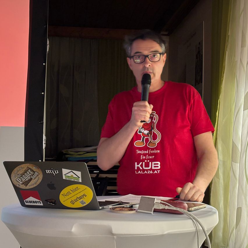 Ein Mann in einem roten T-Shirt mit den Worten KUB LALA26.AT spricht in ein Mikrofon. Vor ihm steht ein Laptop mit verschiedenen Aufklebern. Dahinter eine weiße Gardine und eine Wand mit einem Bild.