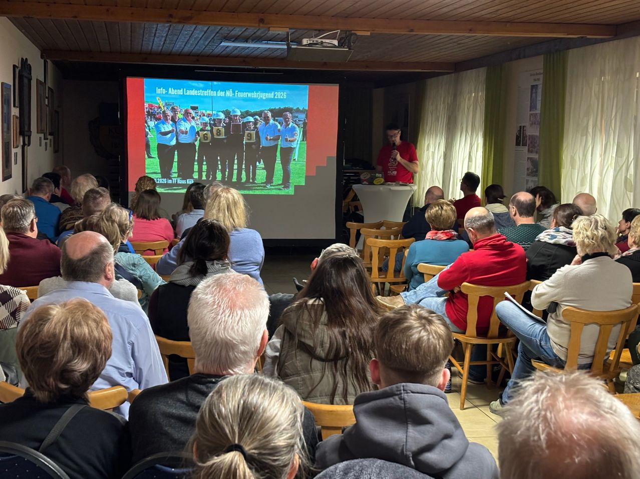 In einem Raum mit Beamer, Rednerpult und Publikum wird eine Präsentation gehalten. Der Moderator hält ein Mikrofon. Die Folie zeigt ein Team von Personen mit Nummern und Text.