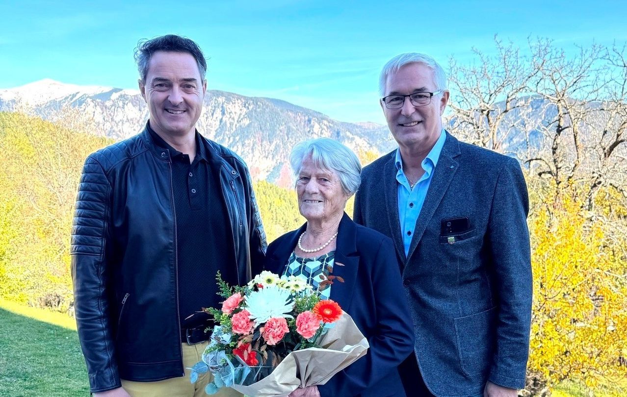 Three individuals, including an elderly woman holding flowers, stand together for a photo with a scenic mountain range in the background.