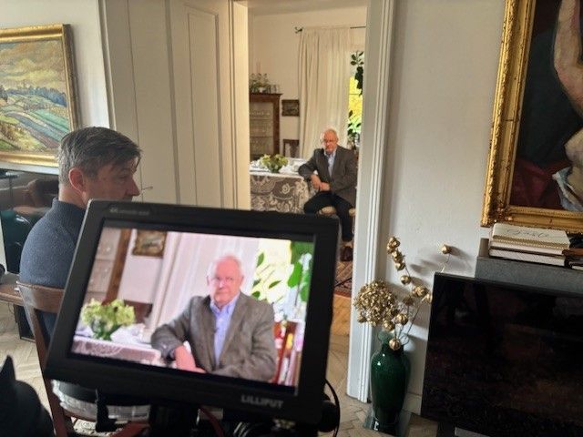 A man is sitting in a chair facing a camera, with another man sitting at a table in the background. A green vase with flowers is on the floor.