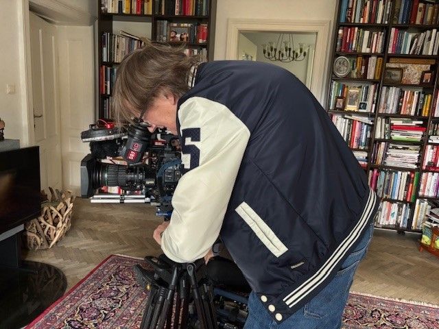 A man in a blue jacket is standing and holding a camera, possibly filming something inside a room with a carpet, bookshelves, and a fireplace.