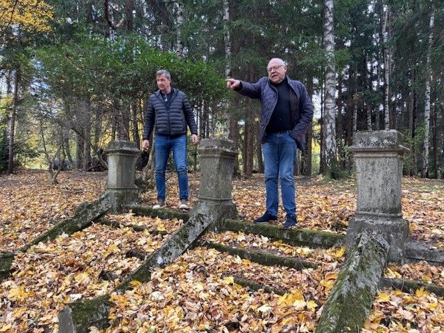 Two men in jackets stand on a fallen concrete structure covered with fallen leaves in a forest. One man points while the other looks on.